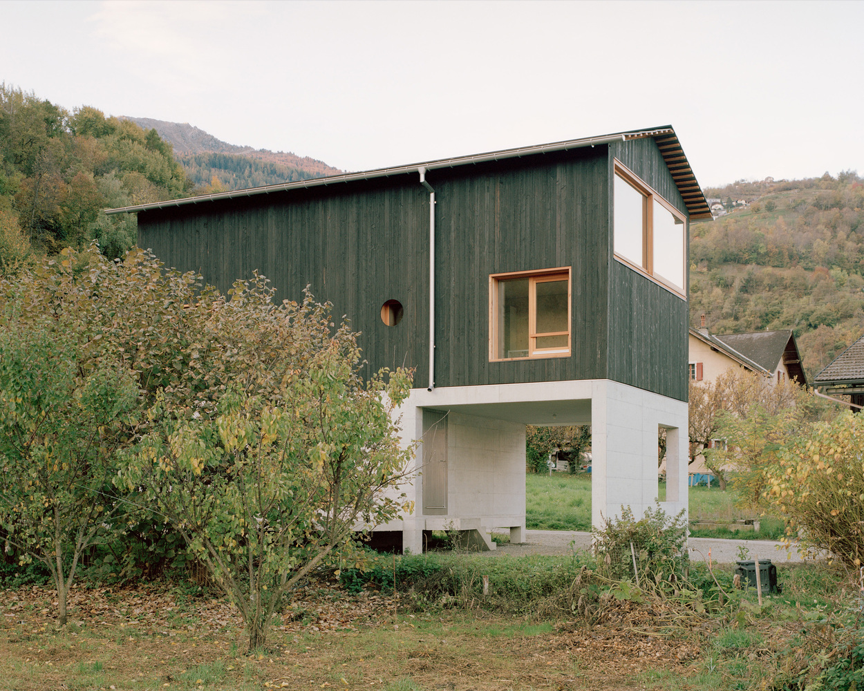 Green vertical timber cladding on the upper volume above a white concrete plinth and covered terrace