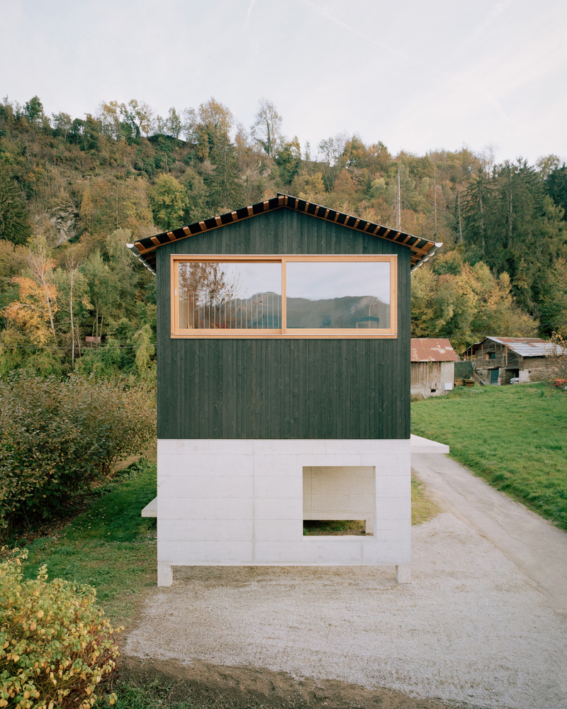 Two-story gable facade with dark timber cladding above a white masonry base and wooded hillside beyond