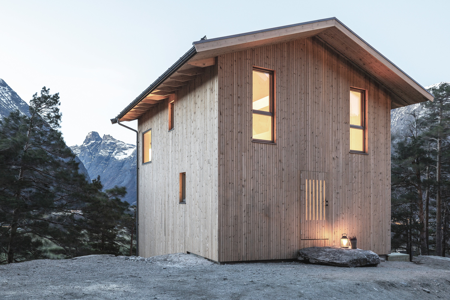 Timber-clad gable facade with illuminated windows at dusk beneath snow-capped mountain peaks