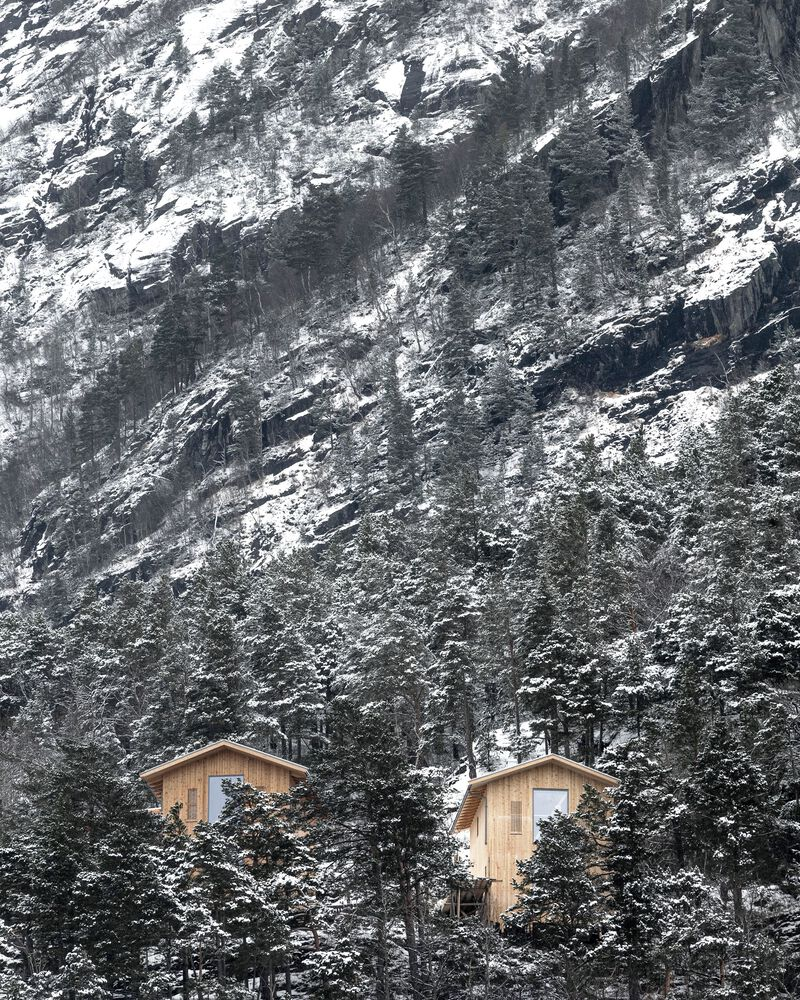 Two timber cabins nestled among pine trees against a steep rocky mountainside dusted with snow