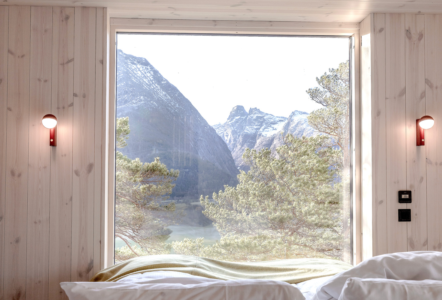 Bedroom window framing snowcapped mountains and riverside trees through light wood paneling with wall sconces