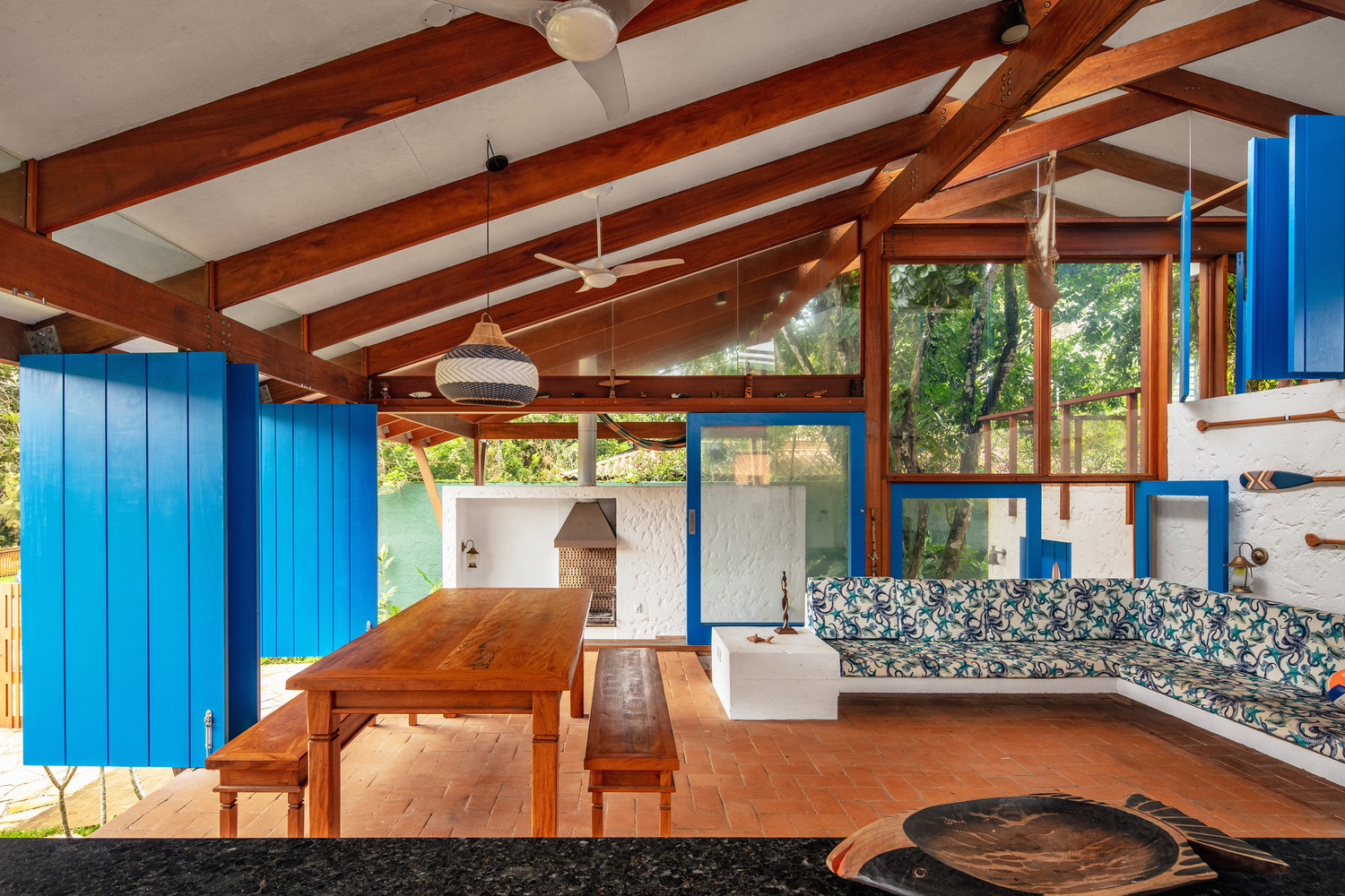 Open living area with timber table, built-in seating, blue partition panels, and views to garden through timber-framed glazing