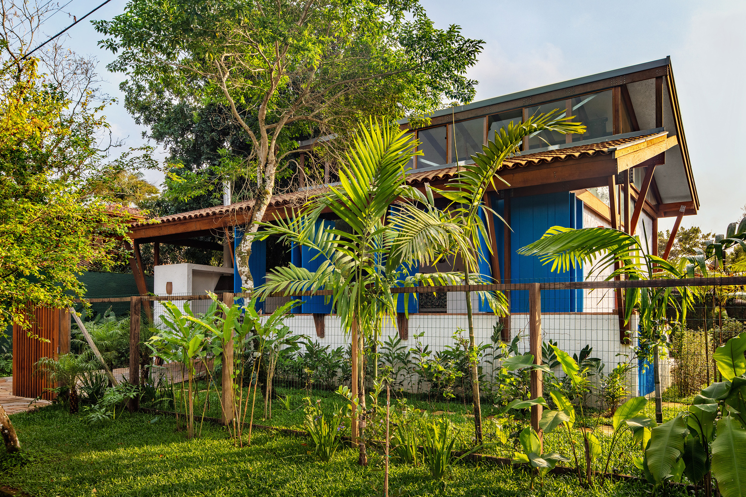 Blue timber-clad volume with glazed upper section and tile roof surrounded by palms and lawn at dusk
