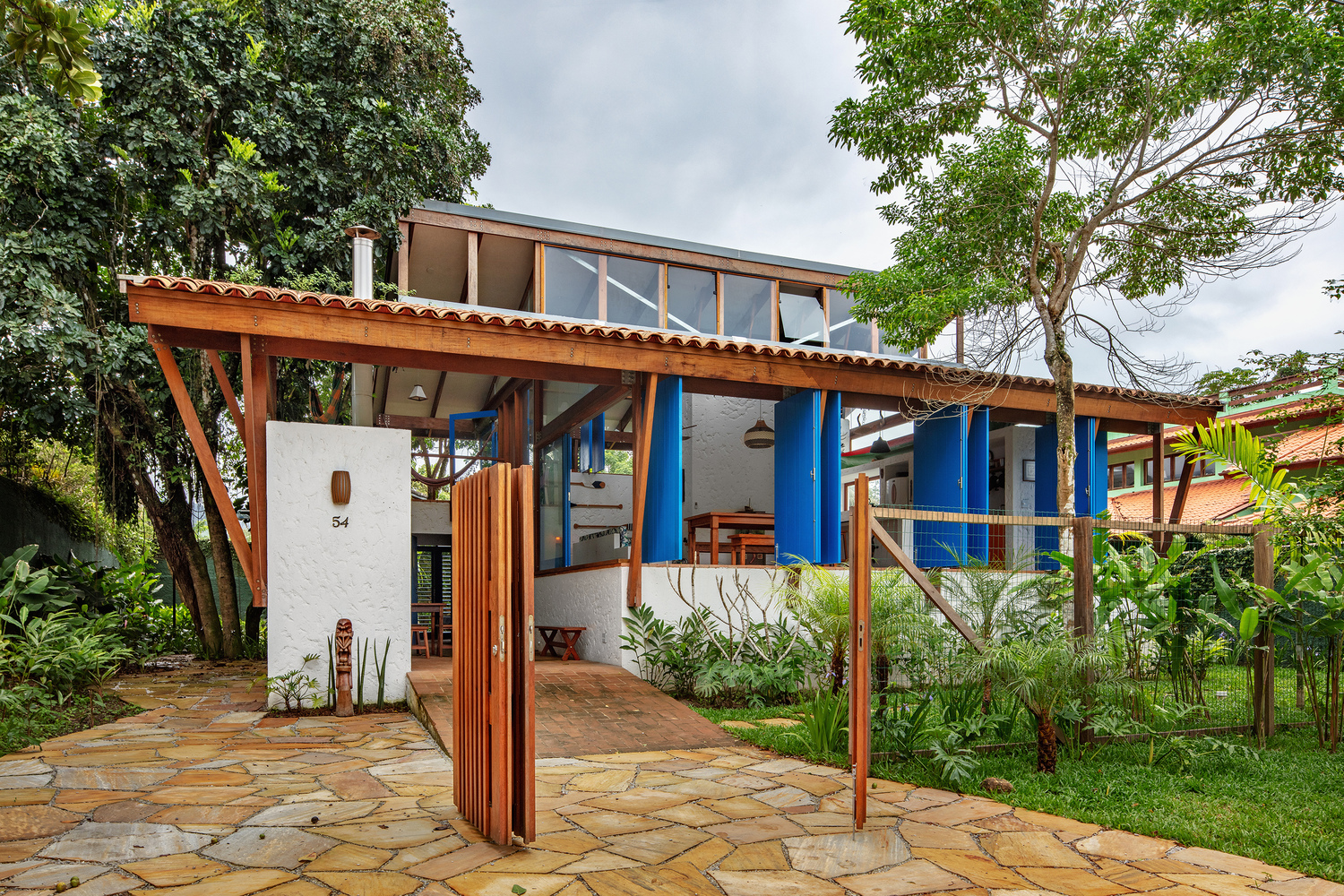 Exterior view of the pitched roof structure with timber trellis, corrugated metal siding, and stone paving amid garden plantings