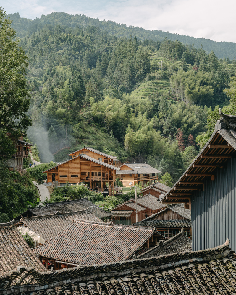 View of timber structures with tiled roofs nestled into a forested hillside under cloudy skies