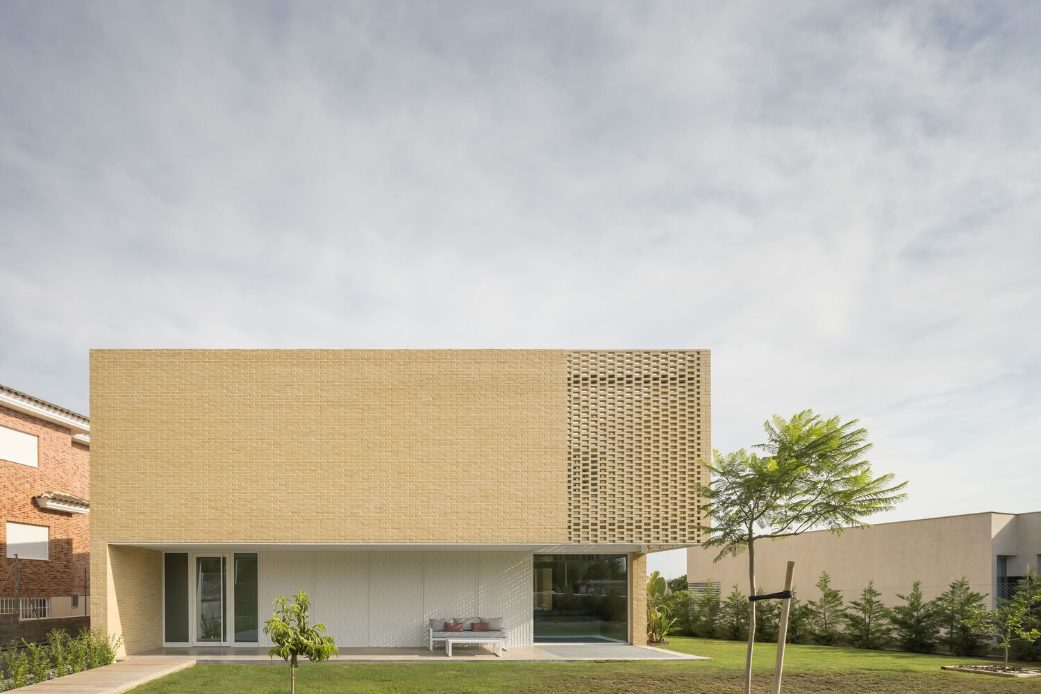 Street view of the pale brick facade with perforated upper volume and lawn with young trees