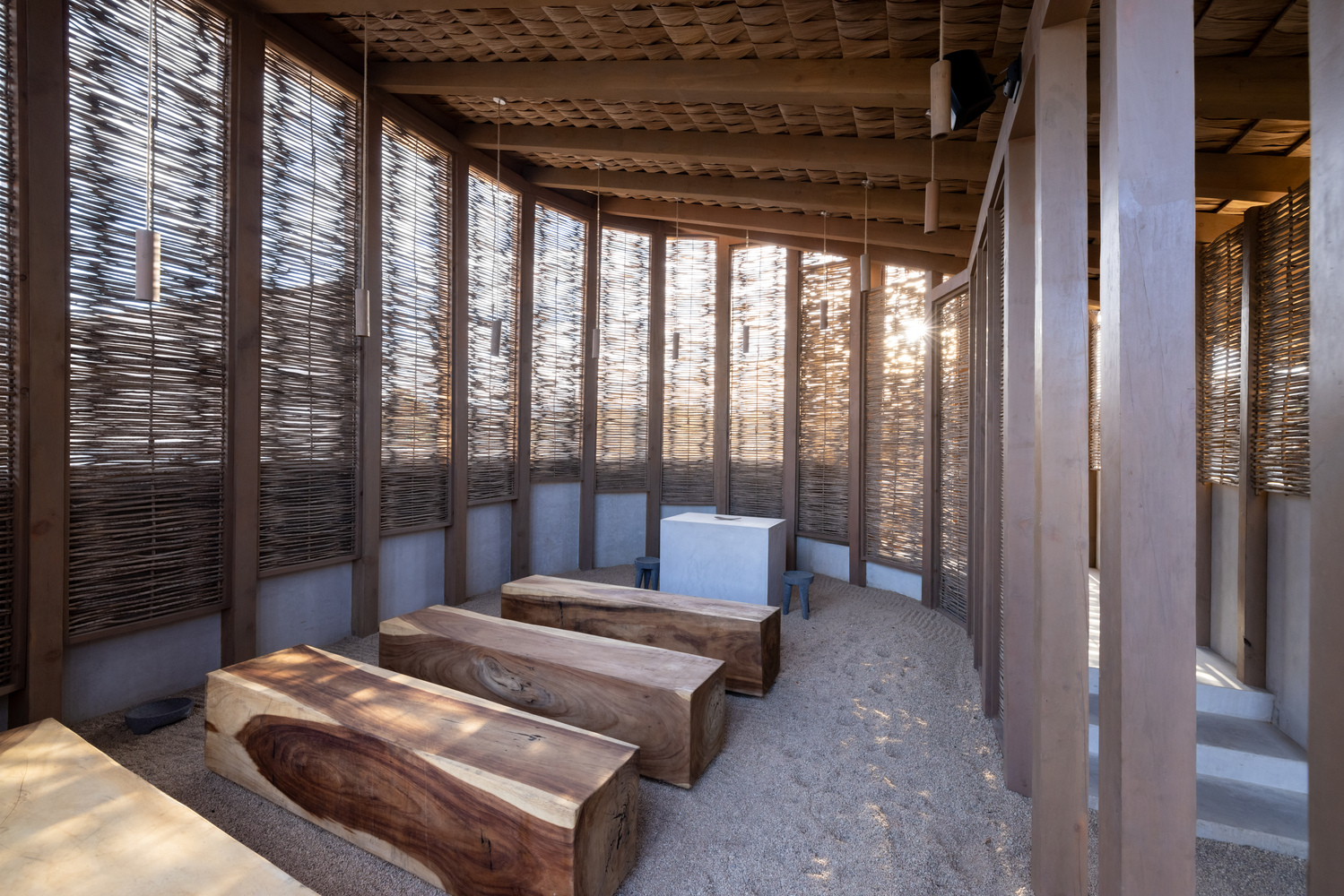 Interior with timber benches on gravel floor beneath exposed rafters and woven screen walls