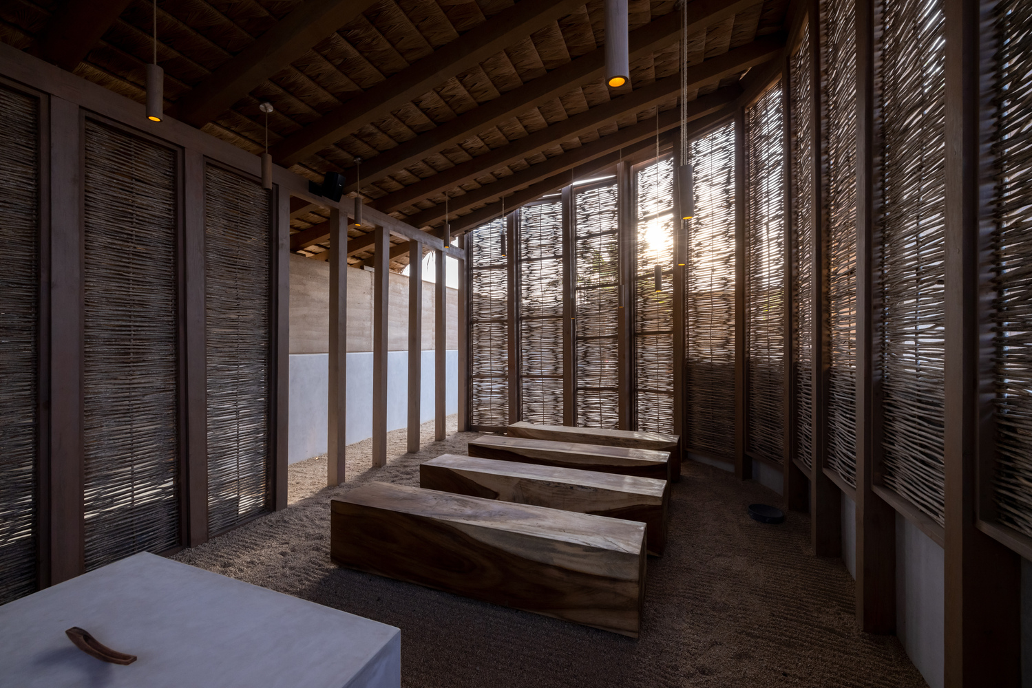Timber benches arranged in rows beneath reed ceiling and backlit woven screen panels at dusk
