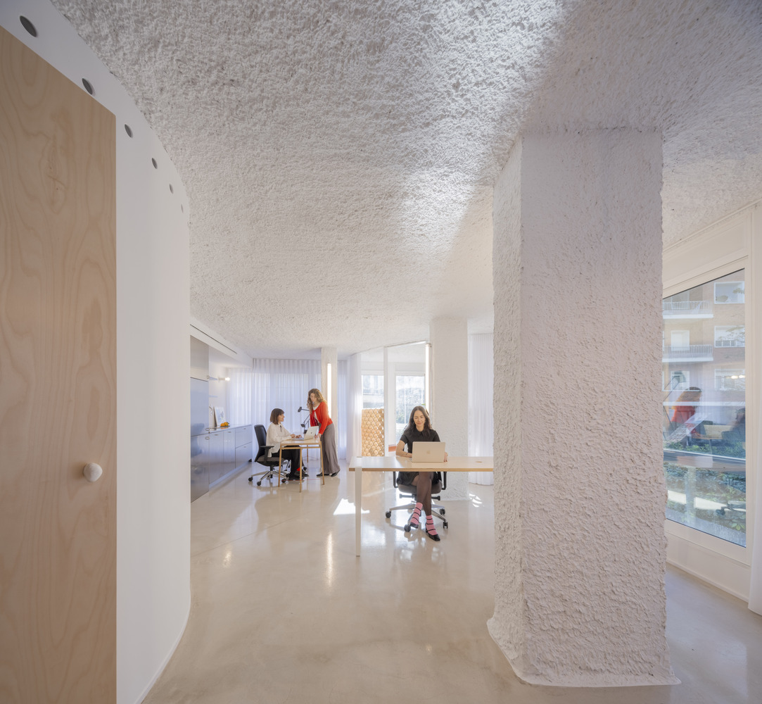 View through textured white columns toward workstations with sheer curtains filtering daylight from windows