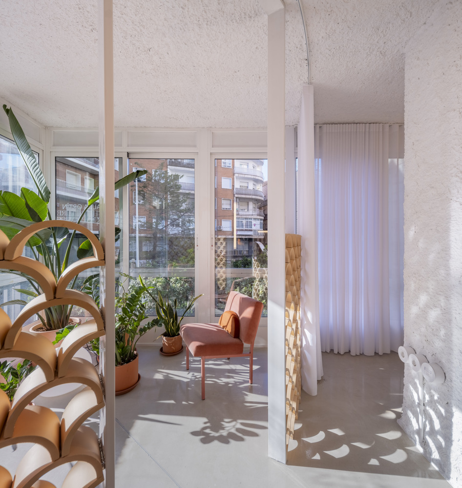 Sunlit seating alcove with potted plants, terracotta armchair, and floor-to-ceiling windows overlooking neighboring buildings