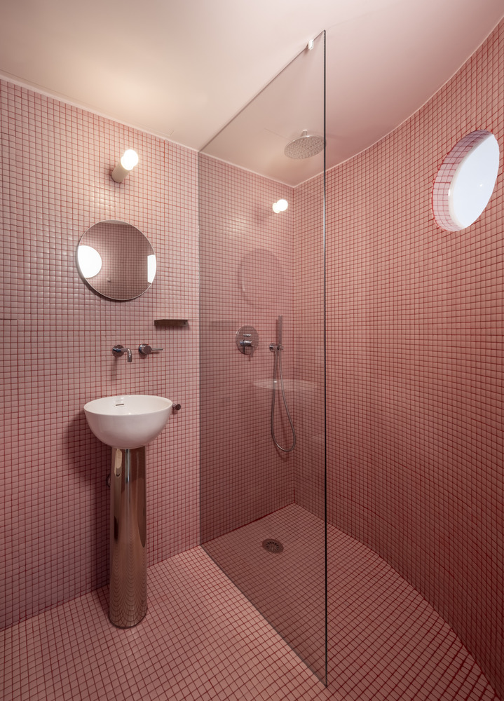 Bathroom clad in pink mosaic tile with pedestal sink, circular mirror, and frameless glass shower partition