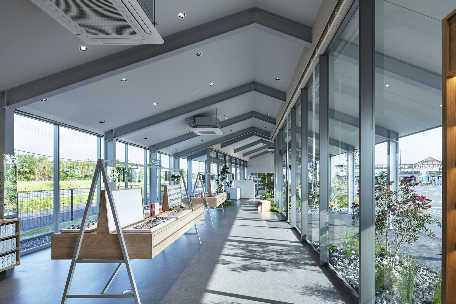 Glass-walled passage with potted trees, display easels, and views to the surrounding landscape