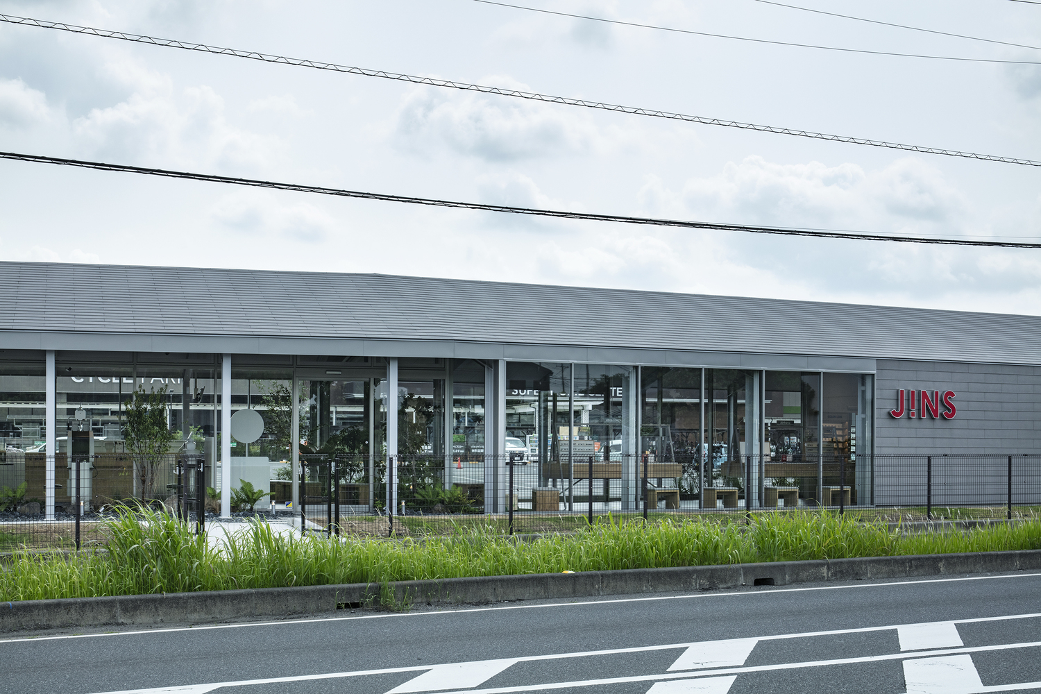 Glazed facade with slender white columns and planted bed along the roadside under overcast sky
