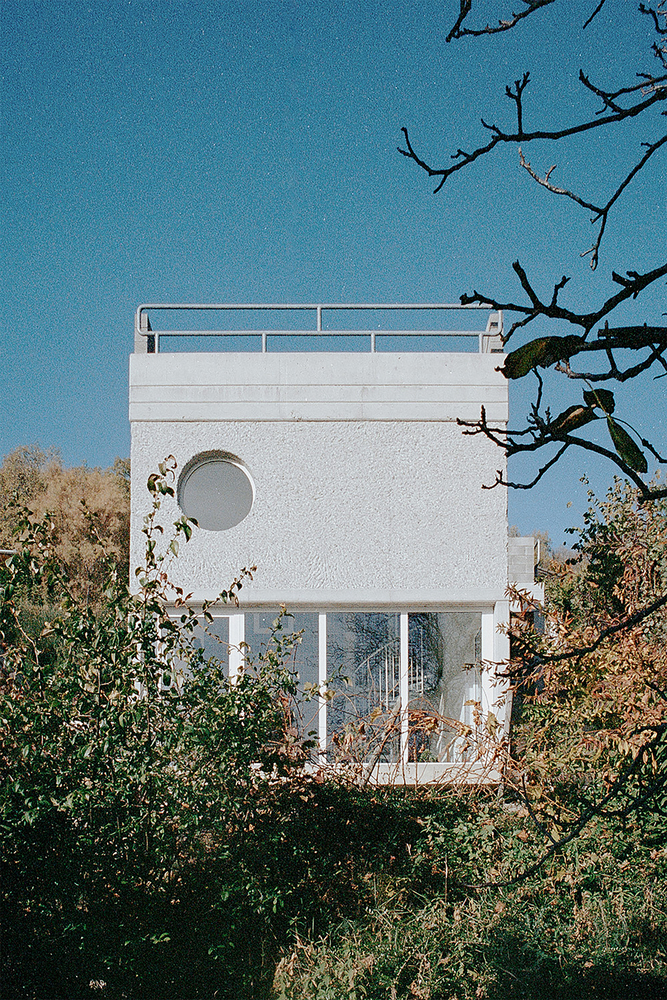 White stucco facade with circular window and rooftop railing framed by overgrown vegetation under clear blue sky