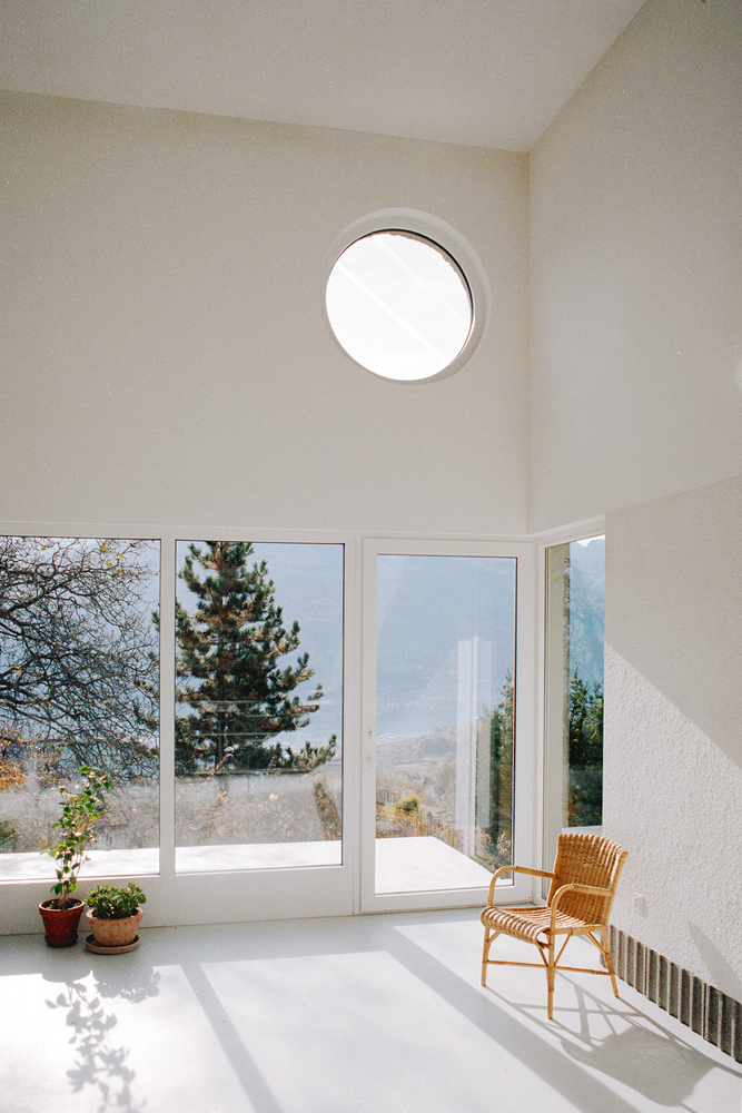 White interior room with circular ceiling window and floor-to-ceiling glazing overlooking hillside with wicker chair in sunlight