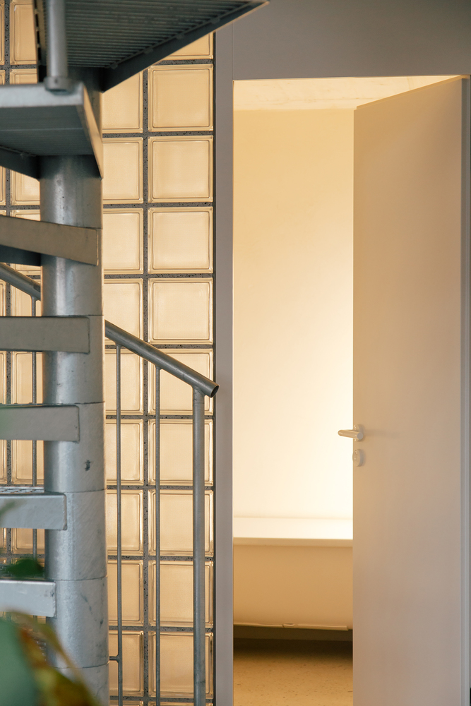 Metal spiral staircase beside translucent glass block wall with warm interior light glowing through
