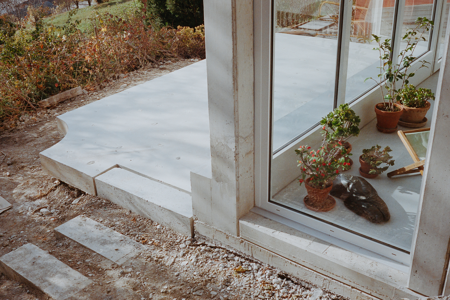 Concrete threshold and gravel bed meeting large window with potted plants on interior ledge at dusk