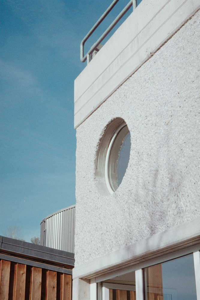 Circular window set into white concrete facade with timber awning and metal railing above under clear sky