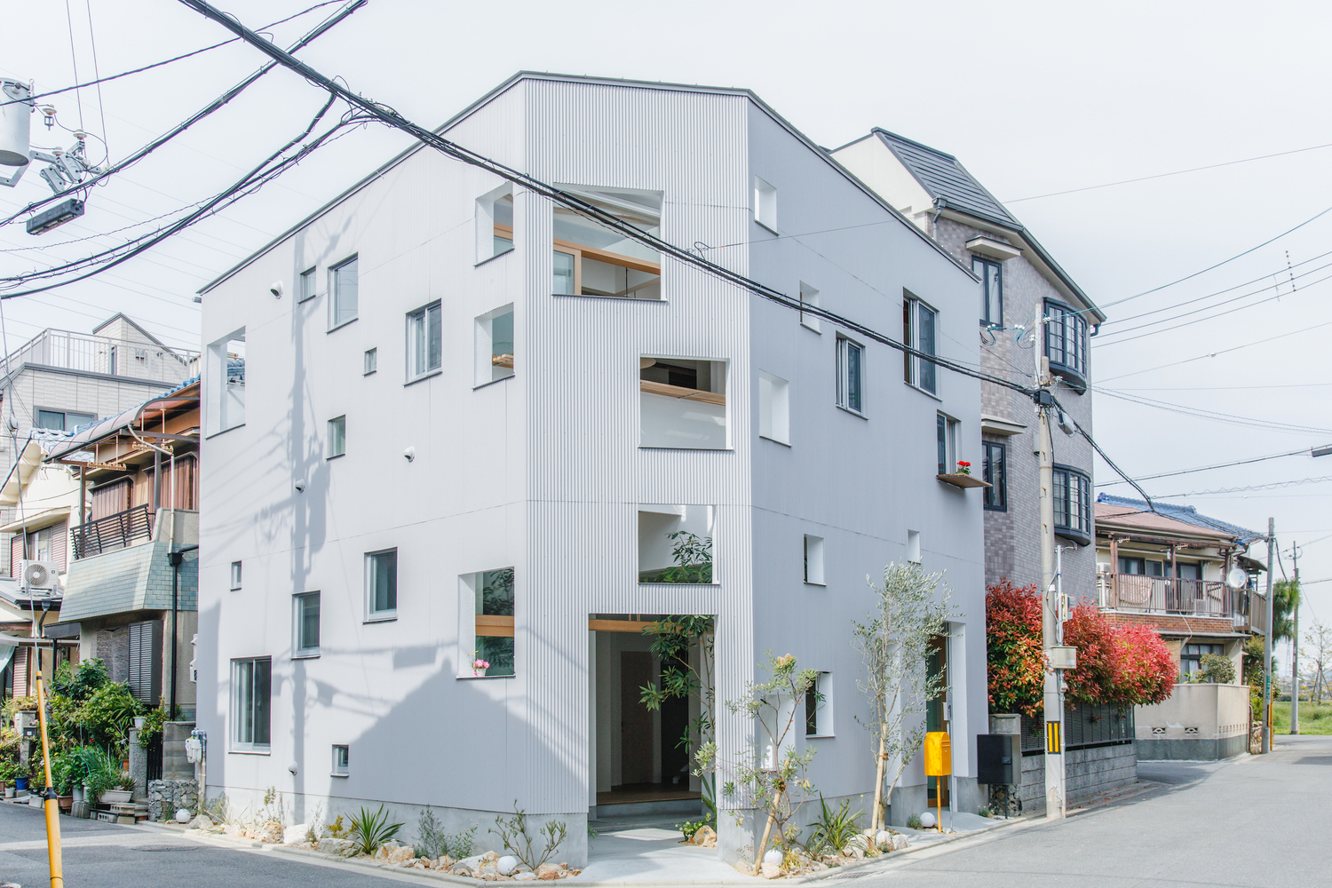 Street view of the white corrugated metal facade with scattered windows and planted front garden