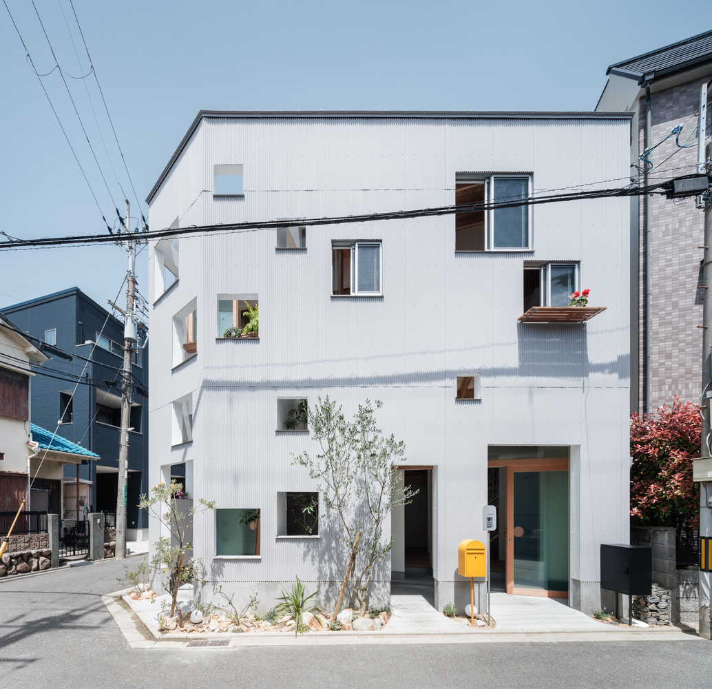 Street elevation showing irregular window openings and young trees in front gravel bed with overhead wires