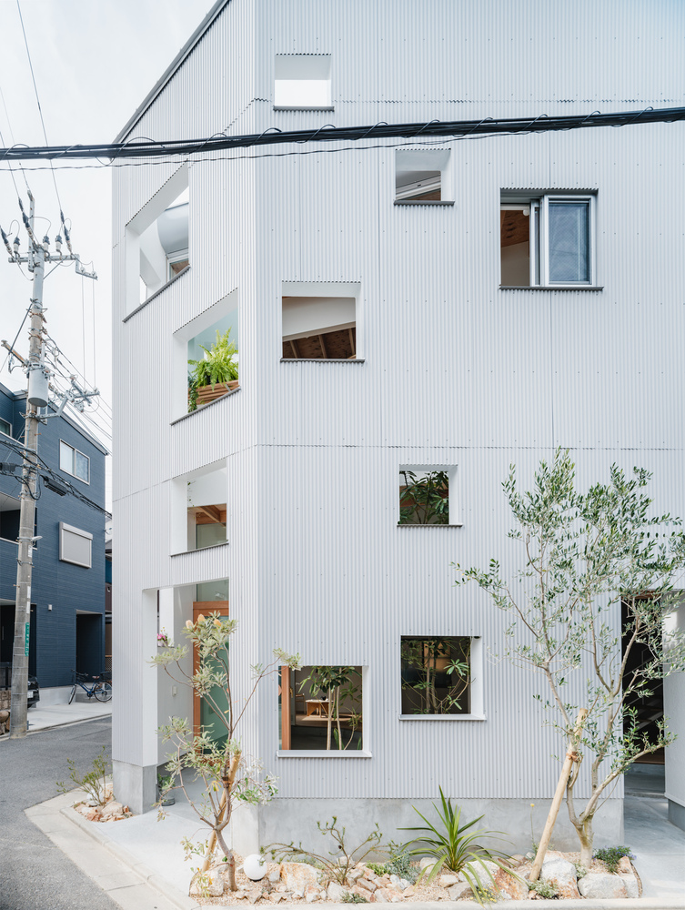 Street view of the white corrugated metal facade with scattered windows and young trees in the front garden