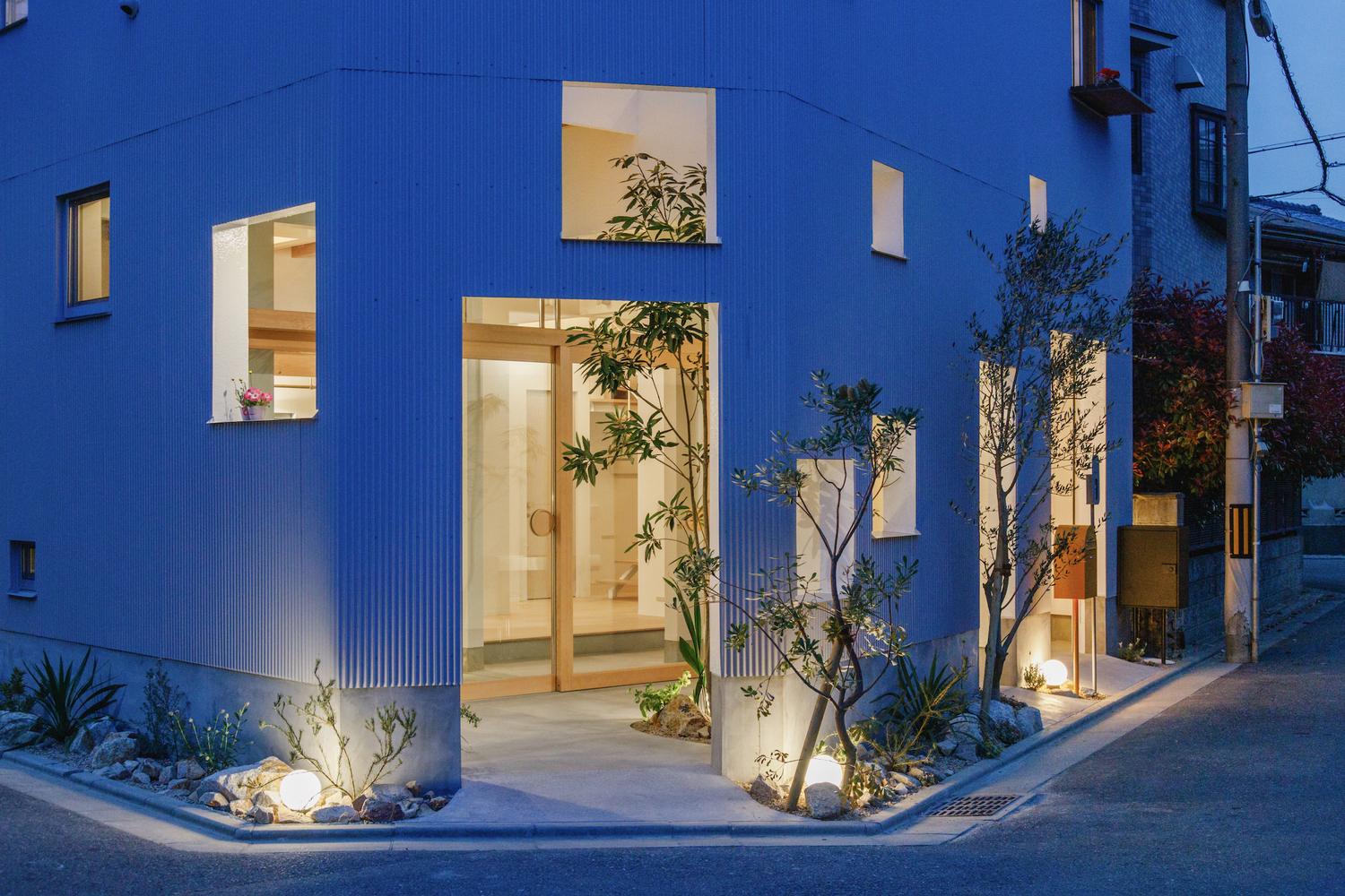 White corrugated metal facade with large glazed openings and uplighting on planted courtyard trees at twilight