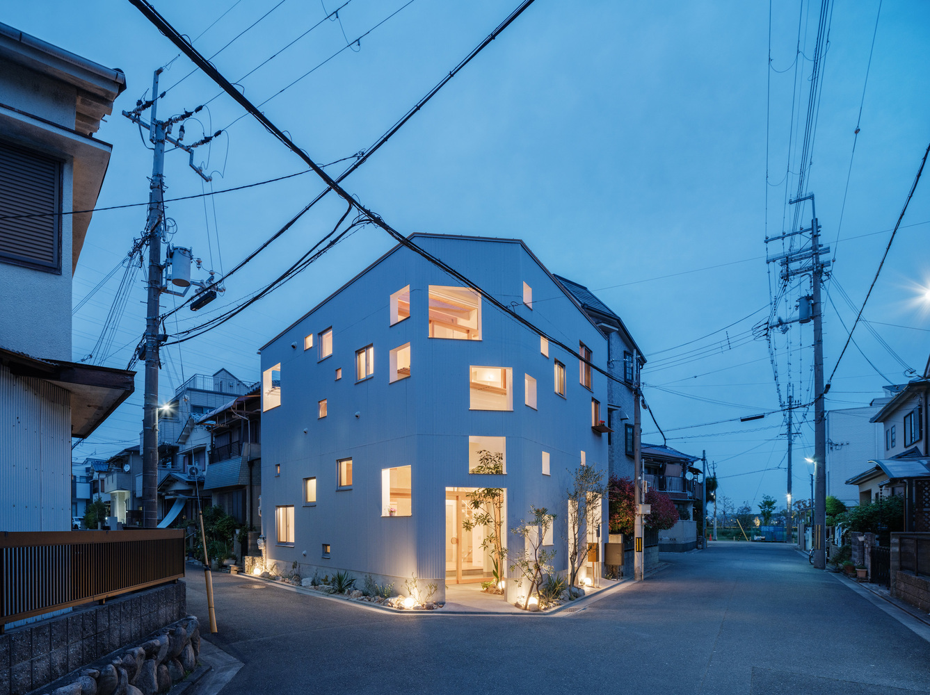 White corner residence with scattered illuminated windows beneath overhead power lines at dusk