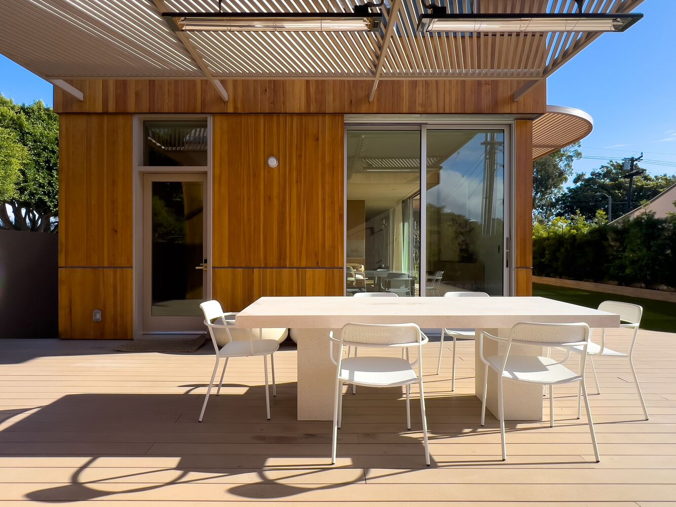 Timber-clad rear facade with slatted pergola overhang and deck dining area under blue sky