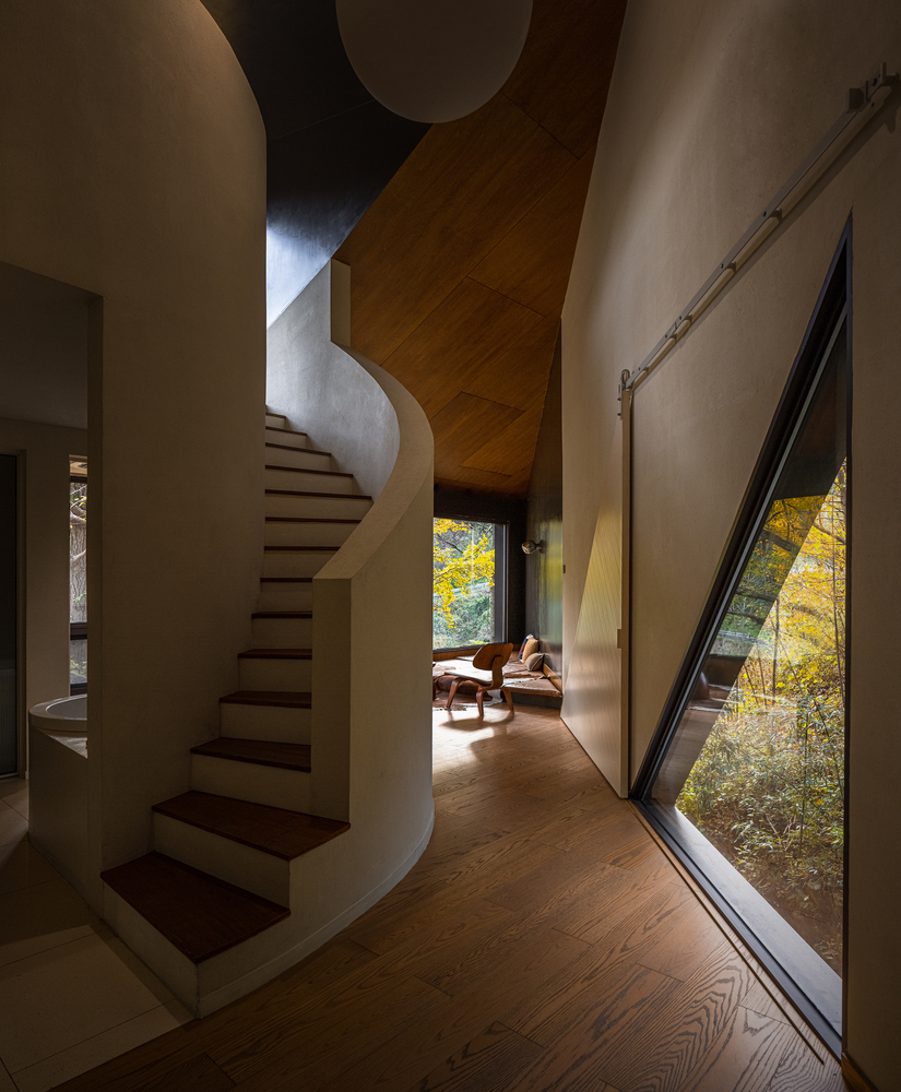 Curved white-plastered staircase ascending beside a large angular window framed in dark timber with autumn foliage beyond
