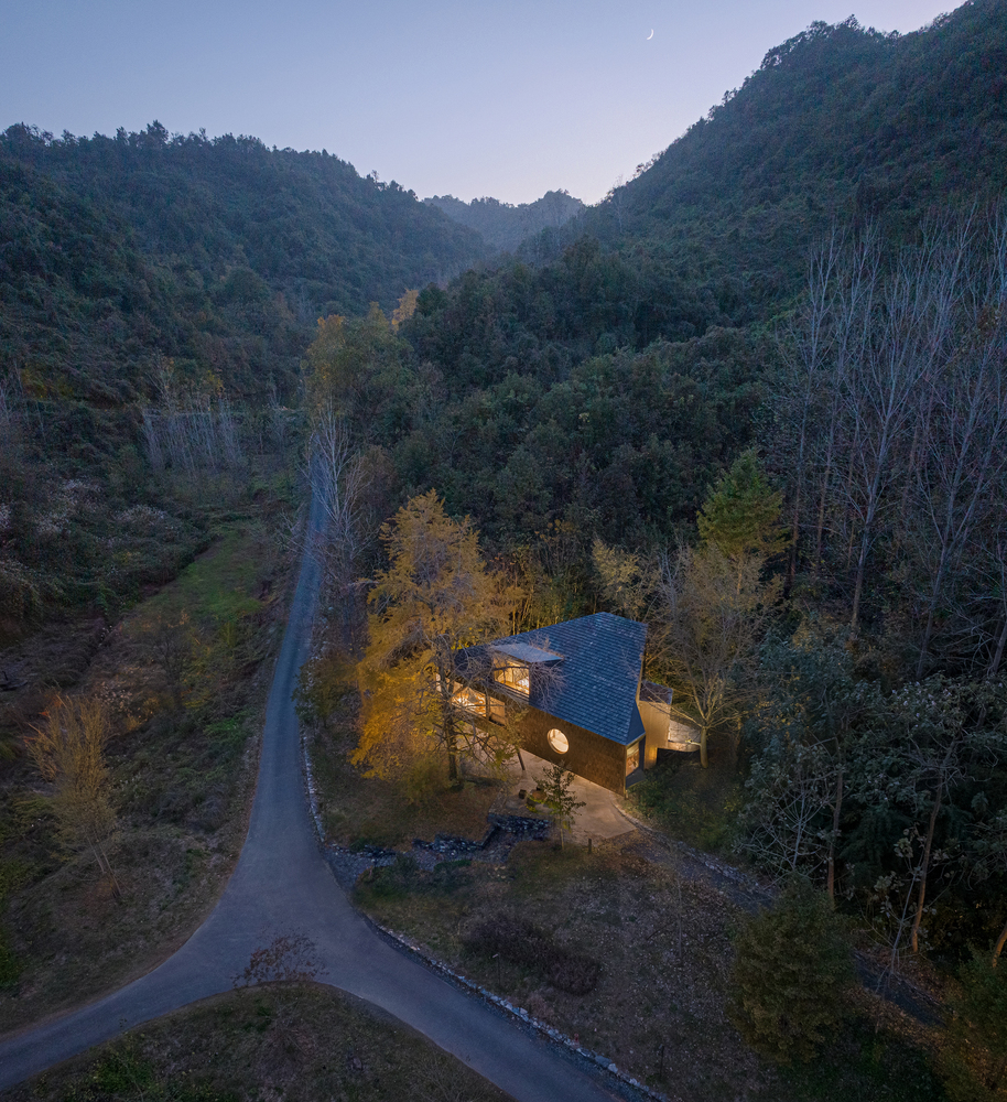 Aerial view of the house nestled among forested hills at dusk with warm interior lighting
