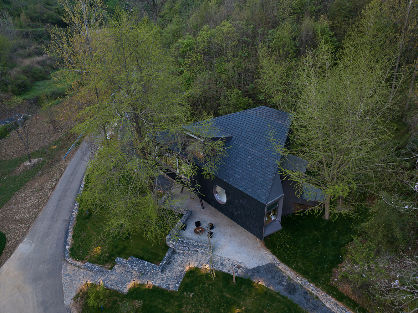 Aerial view of a dark shingled cabin with angled roof nestled among spring trees at dusk