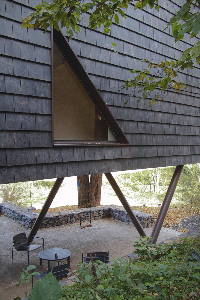 Elevated structure on angled timber supports with dark shingle cladding and triangular window above outdoor seating
