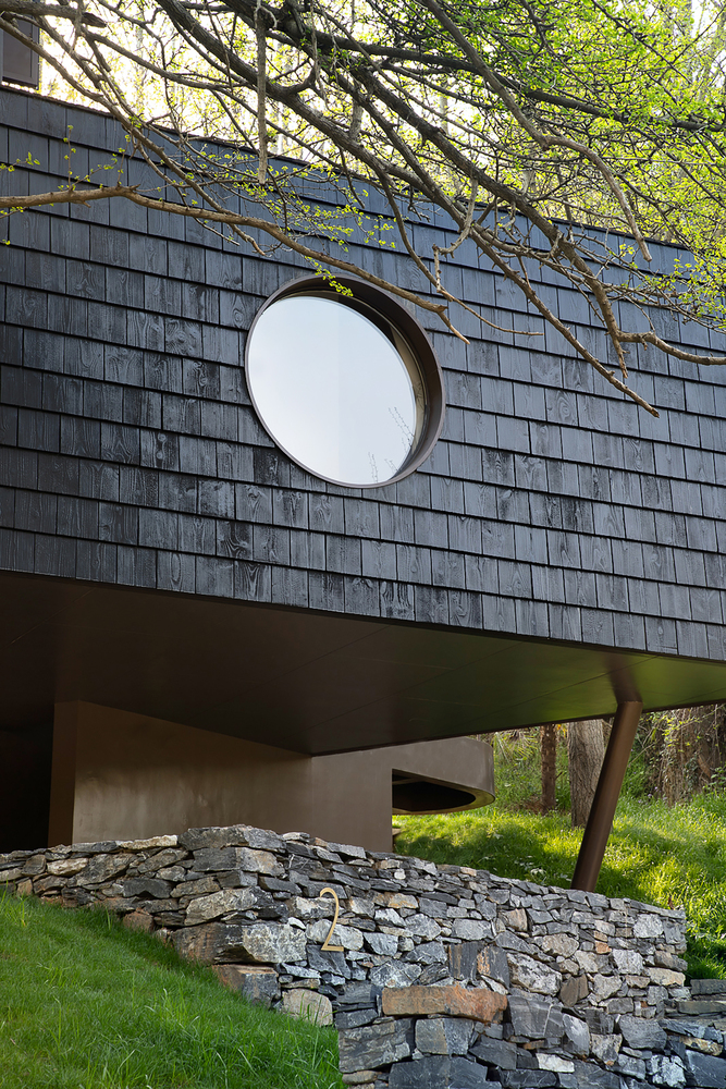 Circular window puncturing dark shingle facade above cantilevered volume and stone retaining wall