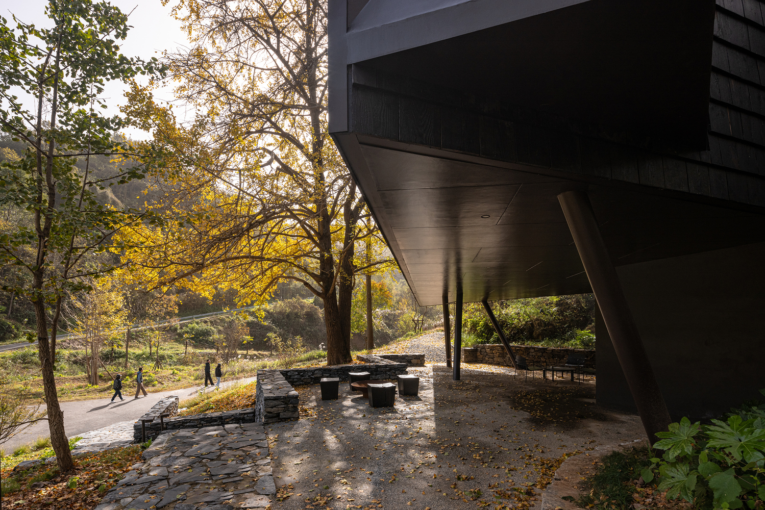 Covered seating area beneath the cantilevered structure with stone pavers and dappled afternoon light