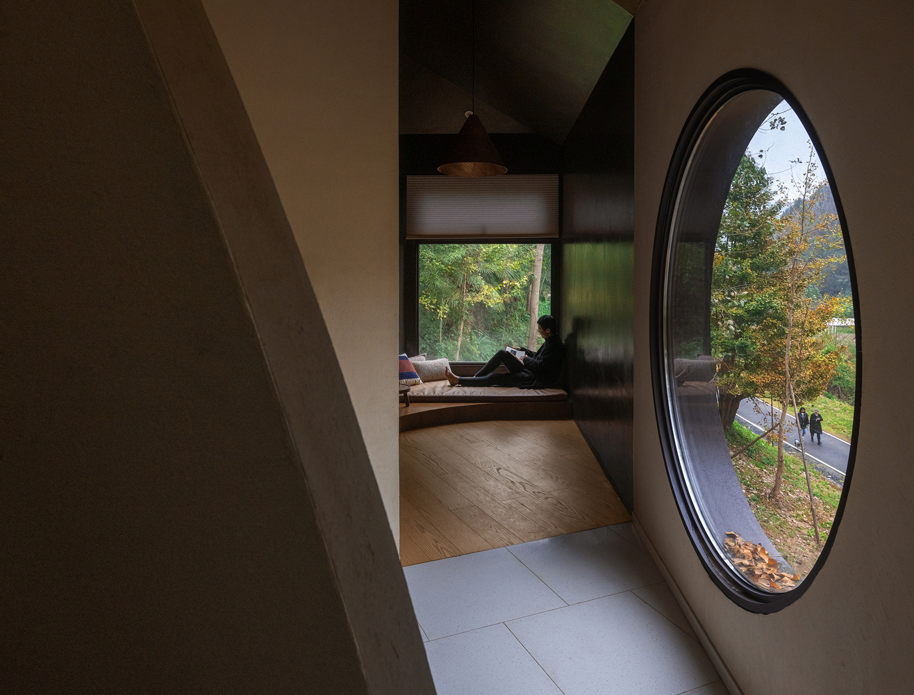 Oval window overlooking the forest path with a figure reclining in the adjacent timber-floored room