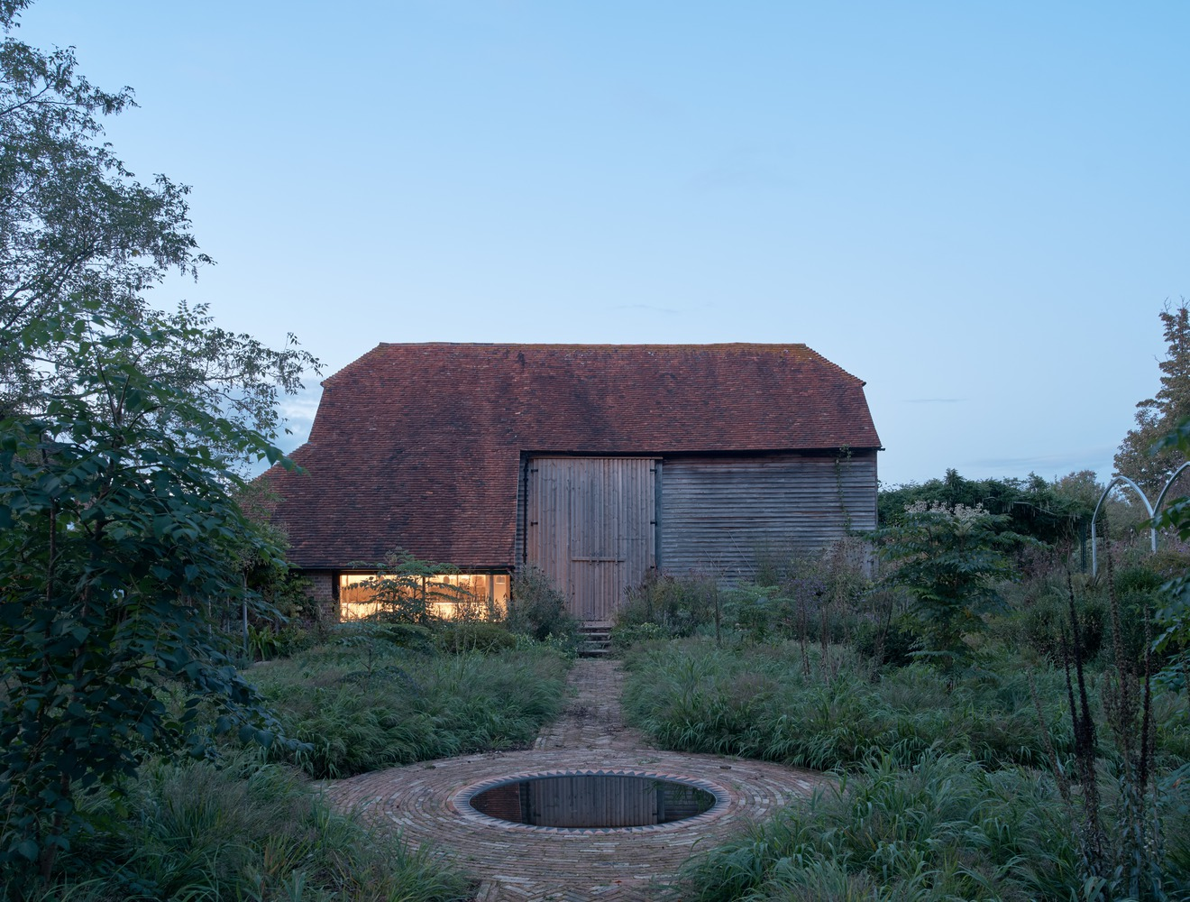 Weathered barn with timber cladding and a circular brick pool in the foreground at dusk