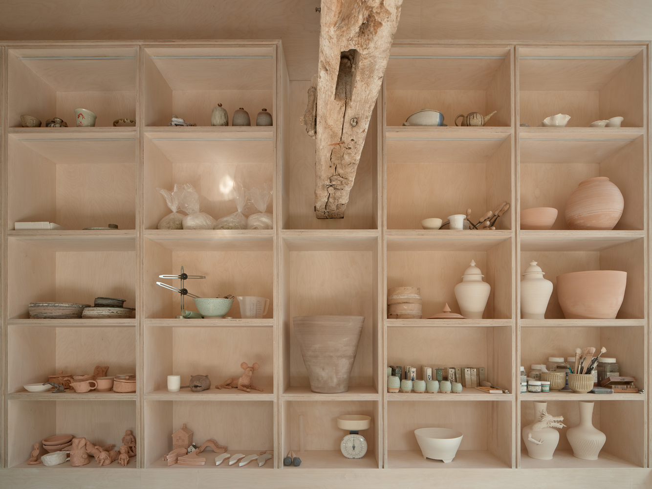 Plywood shelving unit displaying ceramic vessels and materials with a diagonal timber post passing through