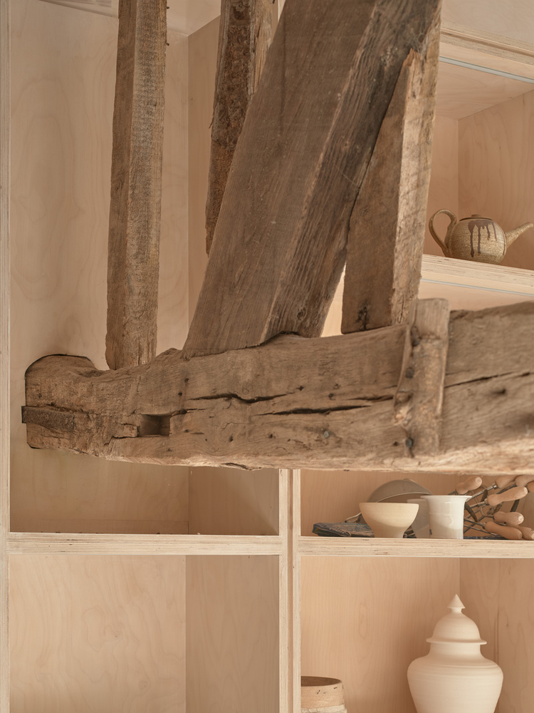 Detail of reclaimed timber beams intersecting above plywood shelving filled with ceramic objects and tools