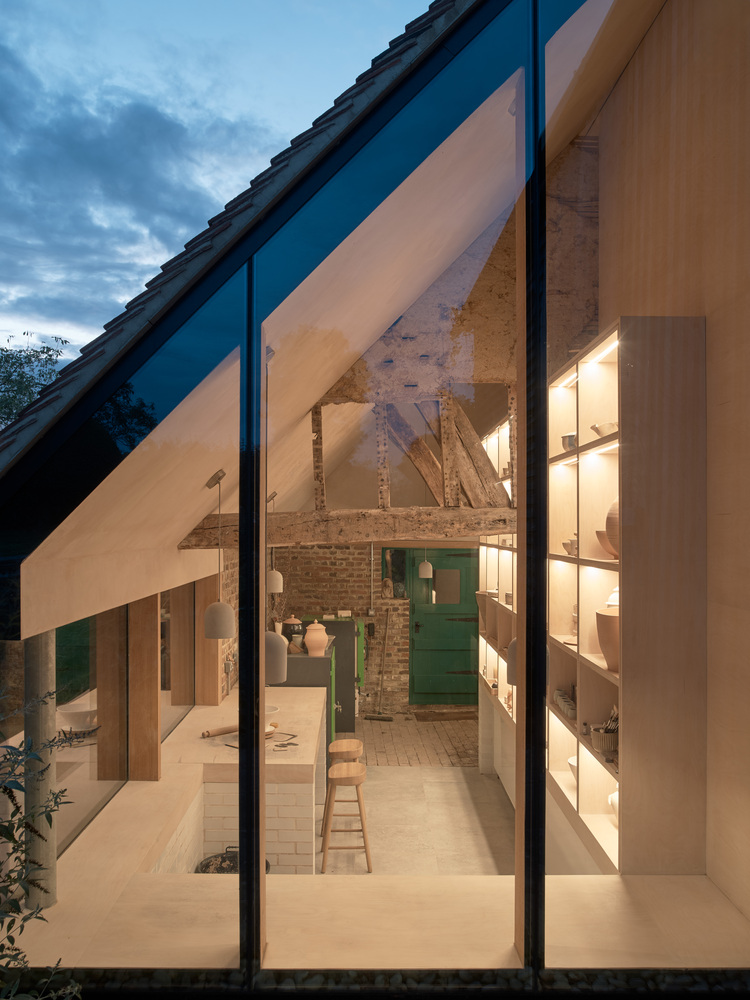 Interior view through full-height glazing showing exposed timber trusses and brick wall at dusk