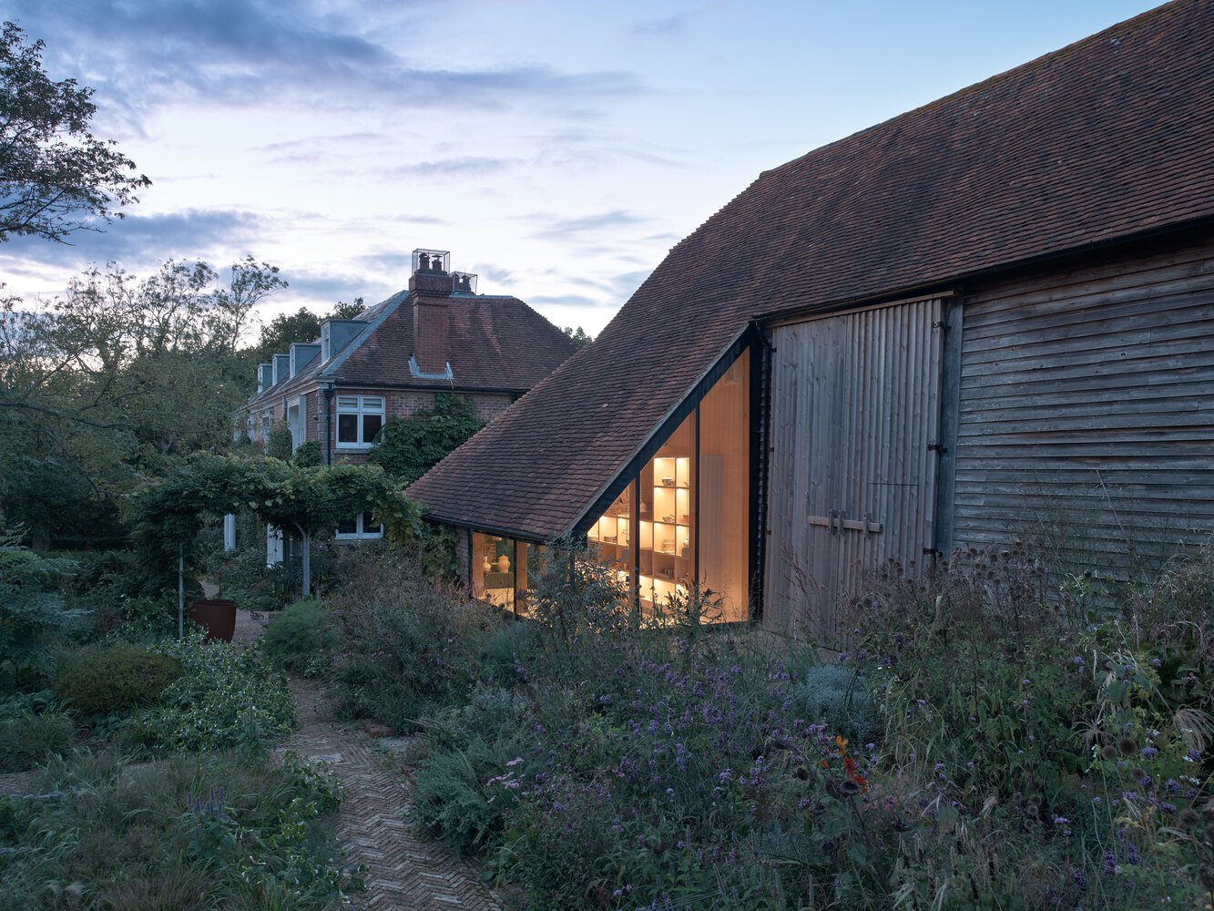 Illuminated timber and glass extension nestled among weathered barns and wildflower garden at dusk