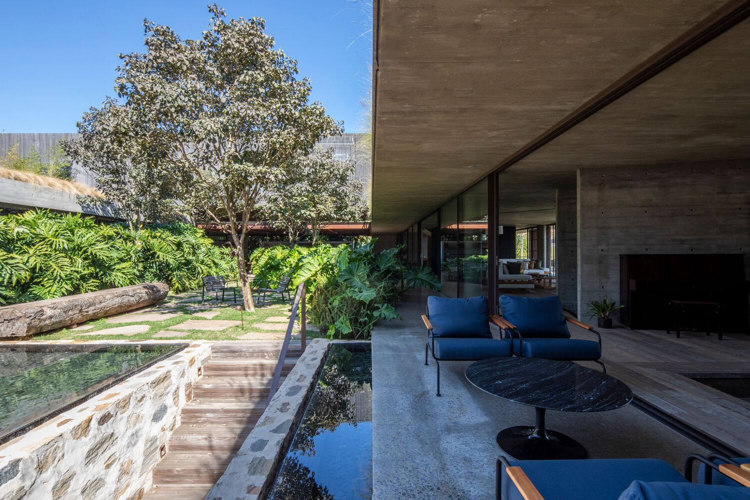 Covered concrete terrace overlooking a courtyard pool with timber bridge and planted stone wall