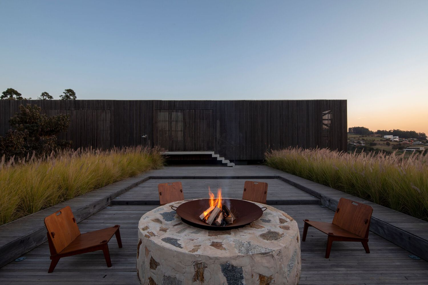 Circular stone fire pit surrounded by timber chairs on a deck at dusk with grasses