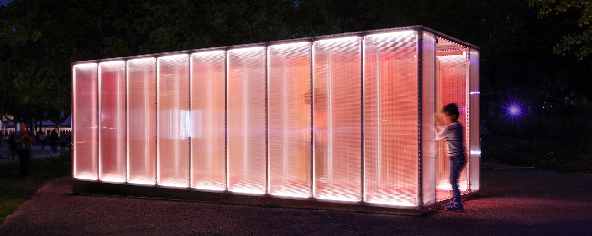 A vibrant illuminated structure stands against the night sky, as visitors interact with its glowing walls, blurring the boundaries of space and light.