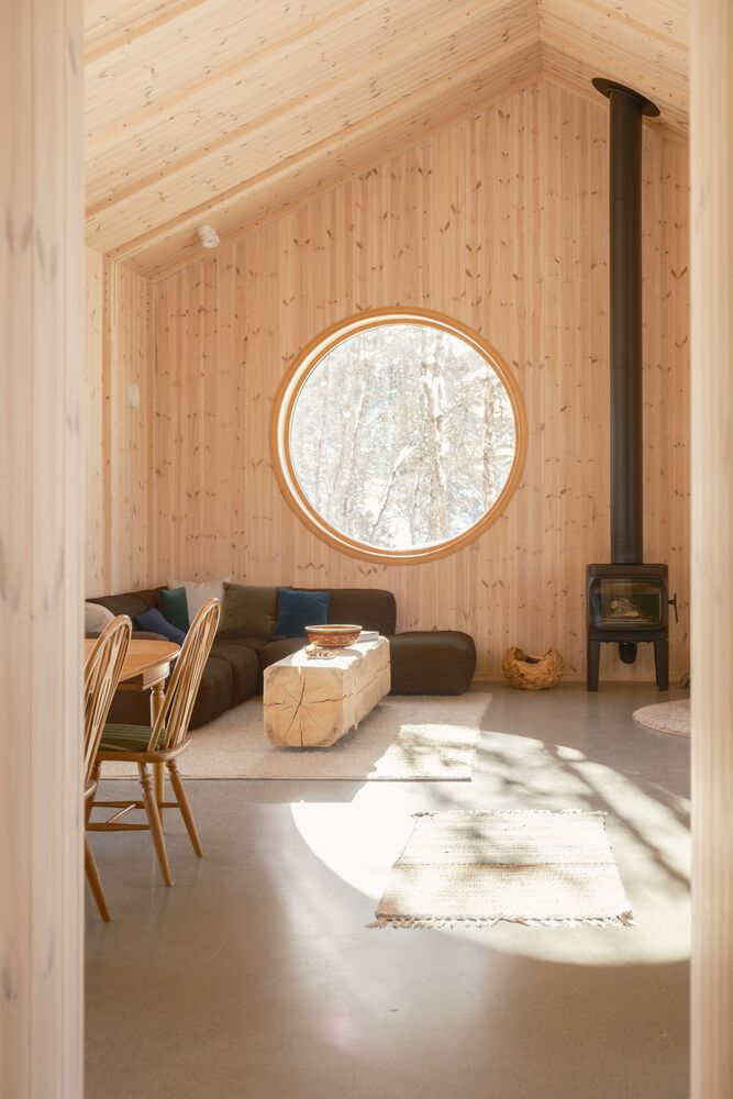 Living area with round window framing birch trees, wood-burning stove and afternoon sunlight across polished concrete floor