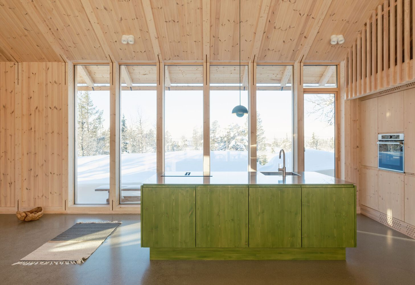 Green kitchen island facing floor-to-ceiling glazing with view of snow-covered birch forest in winter light