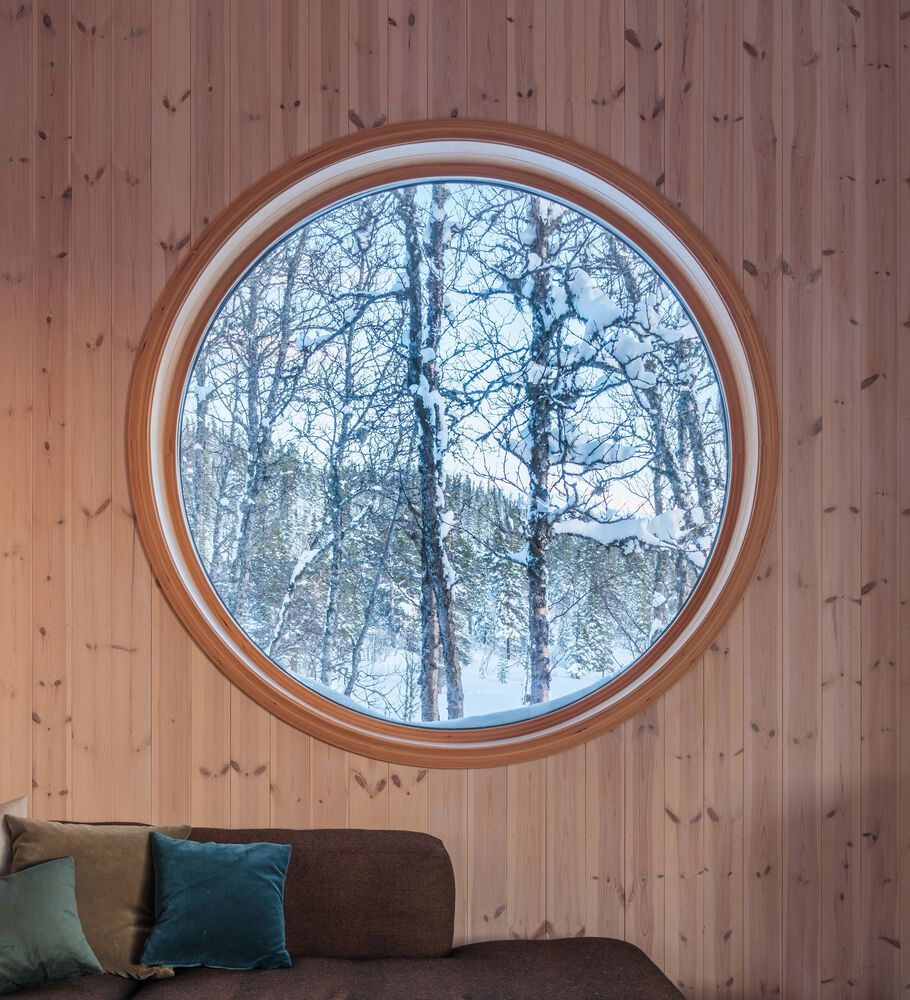 Circular window set in vertical timber paneling framing bare winter trees dusted with snow