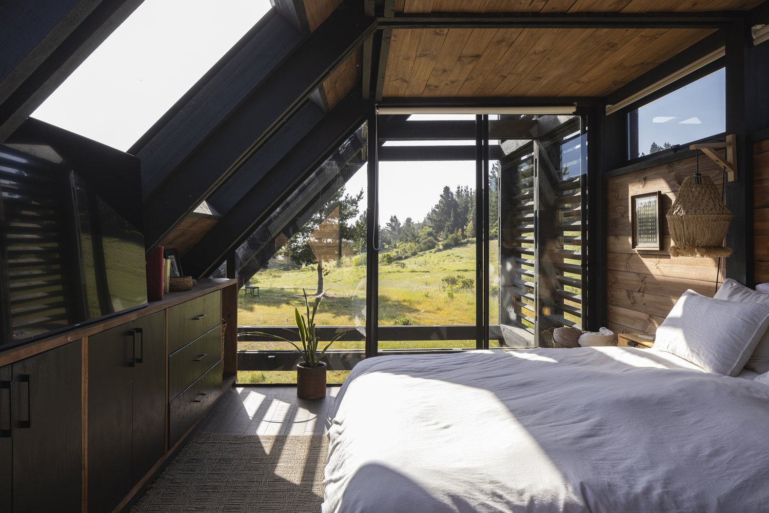 Bedroom with sloped timber ceiling, black steel frame glazing, and morning sunlight across white bedding