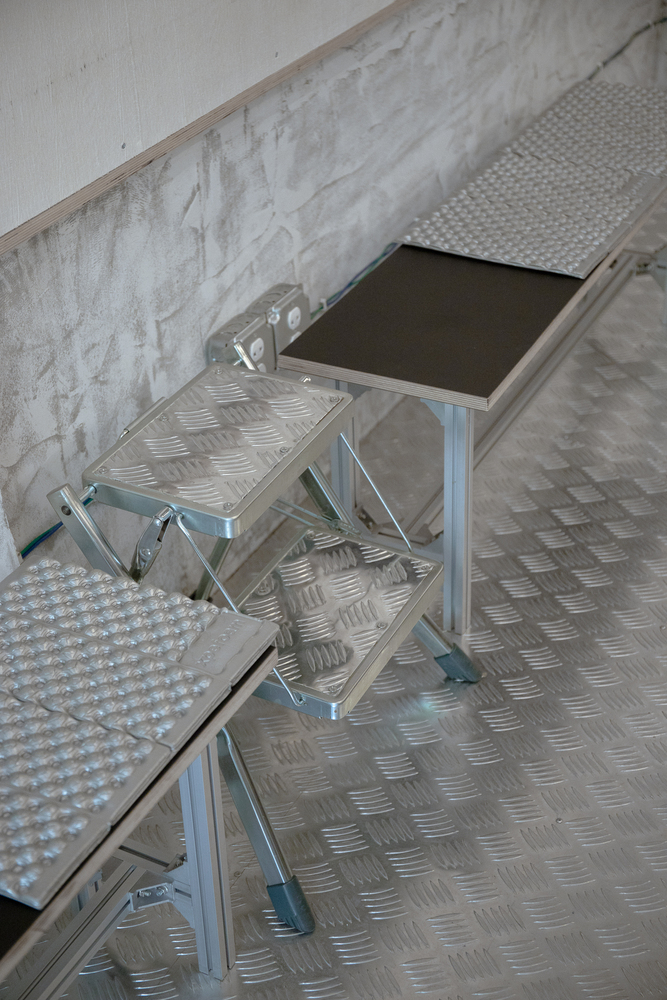 Folding aluminum step ladder on checkerplate metal flooring next to a whitewashed brick wall