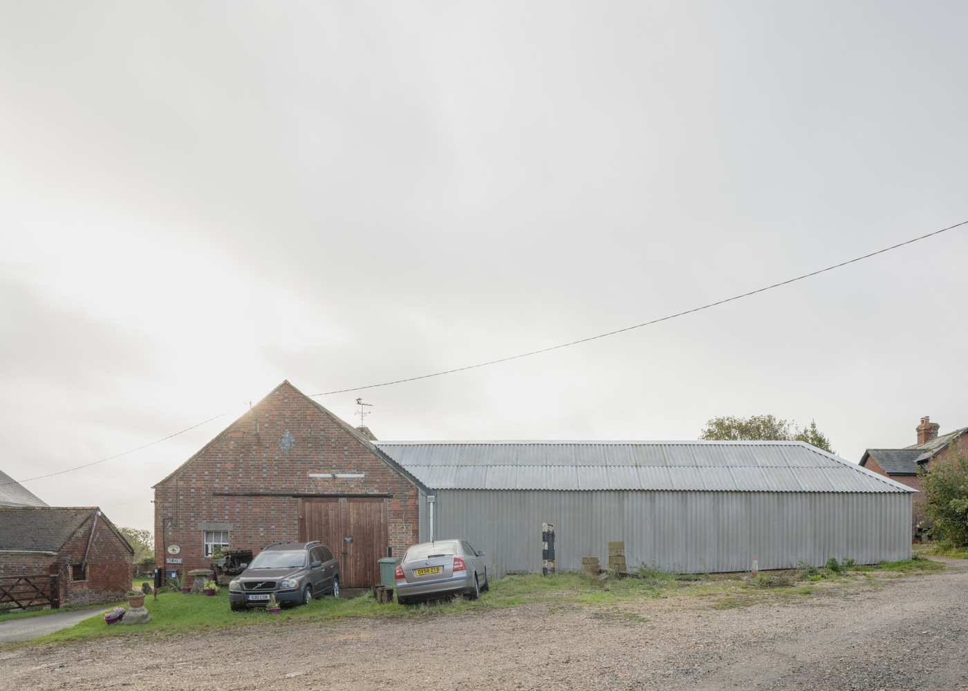 Corrugated metal barn with gabled roof between brick outbuildings and parked cars under an overcast sky