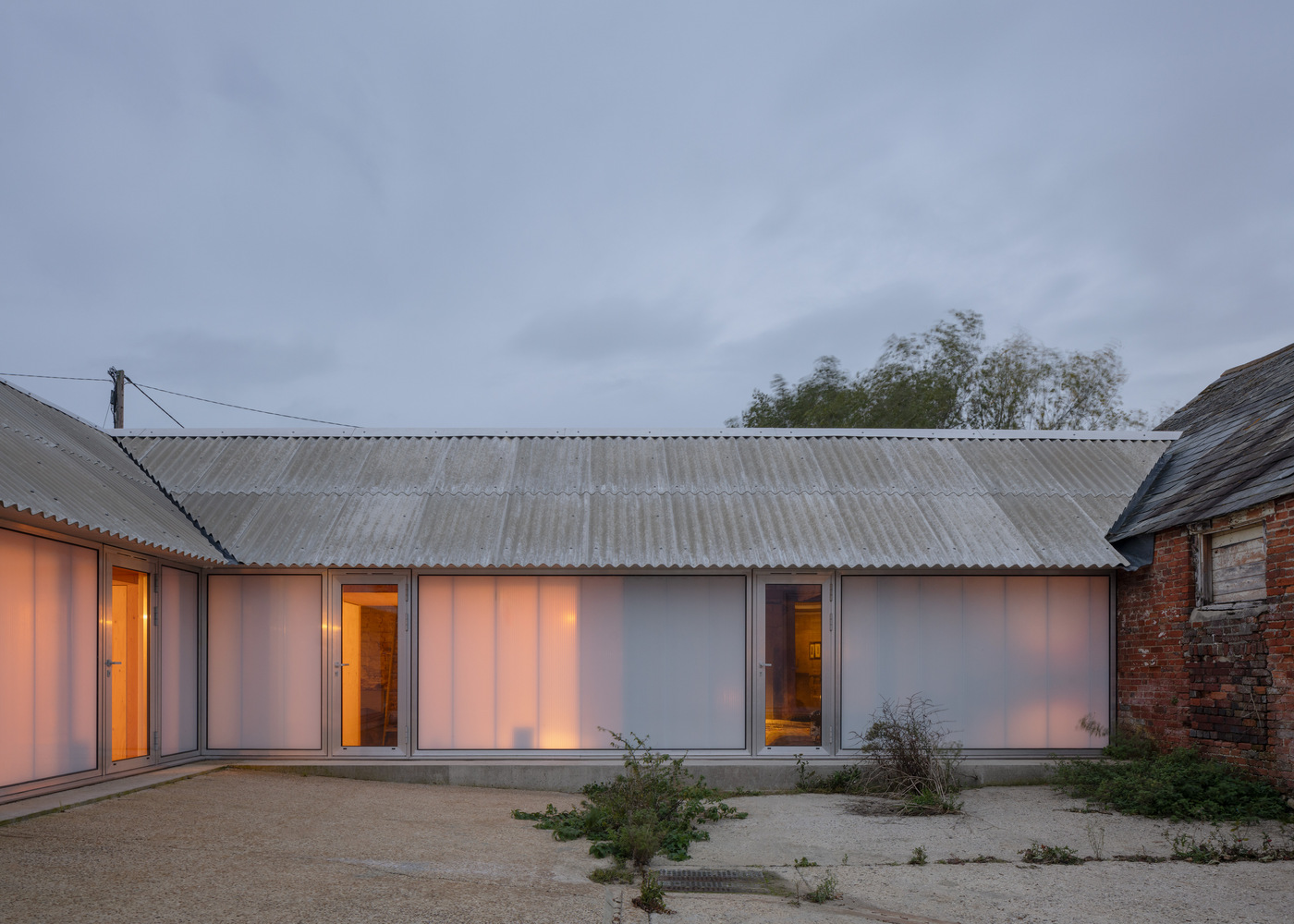 Translucent polycarbonate facade with interior lighting glowing at dusk beneath corrugated metal roof and overcast sky